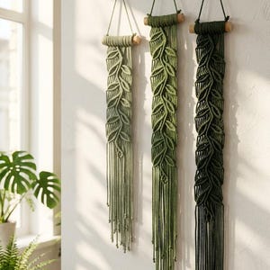 May include: Three macrame wall hangings in varying shades of green, from light sage to dark forest green. Each piece features a leaf-like pattern knotted from cotton cord, suspended from a horizontal wooden dowel with a simple hanging loop.