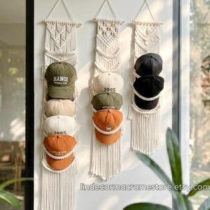 以下が含まれることがあります： 白い壁に掛けられた3つのマクラメハットオーガナイザー。それぞれに野球帽が収納されています。帽子はオリーブグリーン、オレンジ、黒など様々な色があります。オーガナイザーは、下部にフリンジが付いた織りデザインです。