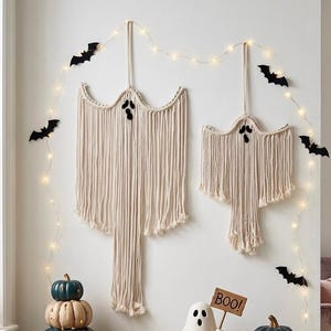 May include: Two off-white macrame ghost wall hangings with black felt eyes and mouths, suspended by cords. The display is accented by a string of warm white fairy lights and several black paper bat cutouts arranged around the ghost decorations.
