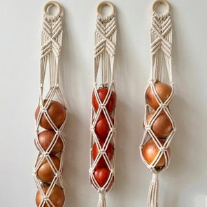 May include: Three cream-coloured macrame hanging holders for fruit and vegetables. Each holder has a wooden ring at the top and a tassel at the bottom. One holds tomatoes, the others hold onions.