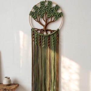 May include: A macrame wall hanging featuring a tree design. The tree has a brown trunk and green leaves within a circular frame. Long, green and brown cords hang below, creating a textured, natural aesthetic. The wall decor is suspended from a loop.