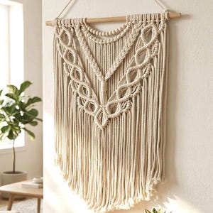 Può includere: Un arazzo da parete in macramè color crema caratterizzato da complessi motivi a nodi e un design a forma di V. L'opera tessile è appesa a un bastone di legno orizzontale tramite una corda, con lunghe frange sciolte sul bordo inferiore.