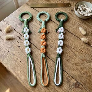 May include: Three macrame keychains featuring circular rings at the top, followed by a vertical row of four crocheted flowers in white or orange, and ending in a long looped cord. The keychains are crafted with green, white, and orange cord.
