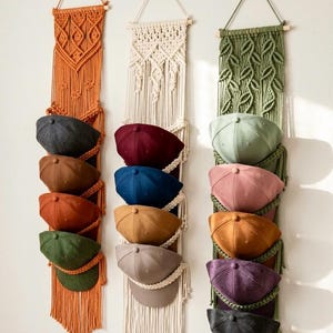 May include: Three macrame hat organizers in orange, cream, and green, each holding several baseball caps. The caps are in various colors, including gray, brown, blue, and green. The organizers are hanging on a white wall.