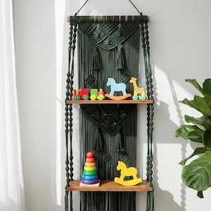 May include: A dark green macrame wall shelf with two wooden shelves. The shelves display colorful wooden toys, including a train, rocking horse, giraffe, and a stacking ring toy. The shelf is suspended by a black cord.