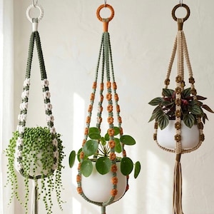 May include: Three macrame plant hangers suspended from hooks, each holding a white ceramic pot. The left hanger is dark green with white flower accents, the centre is sage green with orange flowers, and the right is beige with brown flower details.