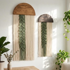 Puede incluir: Dos tapices de pared con partes superiores de madera oscura en forma de semicírculo y largos flecos de algodón color crema. Una tira vertical central de cuerda de macramé verde oliva está anudada en forma de hojas en cada pieza.