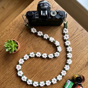 May include: A black 35mm film camera attached to a long, handmade crochet camera strap. The strap features a repeating pattern of white flowers with light pink centers connected by a dark green cord, secured with silver metal swivel clips.