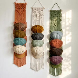 May include: Three macrame hat organizers in shades of orange, cream, and green, each holding five caps. The caps are in various colors, including brown, teal, and beige. The organizers are hanging on a white wall.