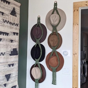 May include: Two macrame hat holders with hats in various colors, including brown, black, and beige, are mounted on a white wall. The holders are olive green and have a tassel at the bottom. A woven wall hanging is on the left.