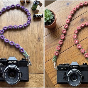 May include: Two black Canon FTb film cameras, each attached to a handmade crochet camera strap. One strap features purple flowers with green centers, while the other features pink flowers with white centers, both connected by silver metal clips.