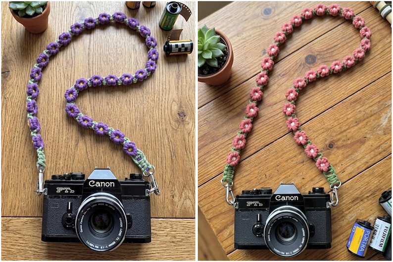 May include: Two black Canon FTb film cameras, each attached to a handmade crochet camera strap. One strap features purple flowers with green centers, while the other features pink flowers with white centers, both connected by silver metal clips.