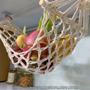 May include: A cream macrame fruit hammock, filled with colourful fruit including dragon fruit. The hammock is suspended from a wooden dowel and is designed to hang from the ceiling. The Etsy shop name is visible at the bottom.