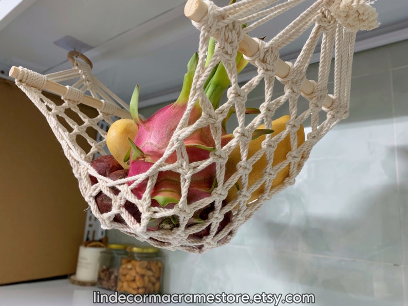 May include: A cream macrame fruit hammock, filled with colourful fruit including dragon fruit. The hammock is suspended from a wooden dowel and is designed to hang from the ceiling. The Etsy shop name is visible at the bottom.