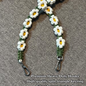 May include: A camera strap featuring a series of crocheted white daisy flowers with yellow centers attached to a dark green cord. The strap includes two silver-toned metal swivel hooks at the ends. Text reads: Premium Heavy Duty Hooks, High quality split triangle keyring, Fit and carry easily all supports cameras on the market!