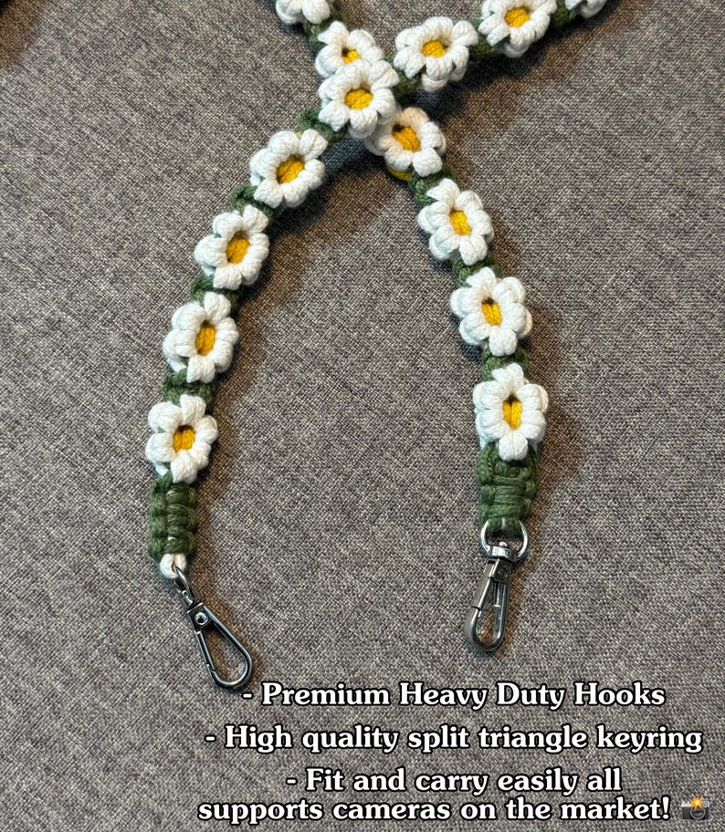 May include: A camera strap featuring a series of crocheted white daisy flowers with yellow centers attached to a dark green cord. The strap includes two silver-toned metal swivel hooks at the ends. Text reads: Premium Heavy Duty Hooks, High quality split triangle keyring, Fit and carry easily all supports cameras on the market!