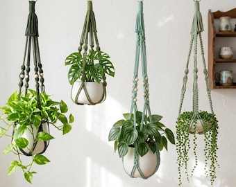 May include: Four hanging macrame plant holders in shades of green and blue-green, each holding a different type of leafy green plant in a white ceramic pot. The macrame hangers have wooden bead accents and are suspended from metal hooks.