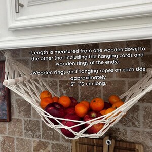 May include: A white macrame fruit hammock, filled with red apples and oranges, suspended from a wooden dowel. The text on the image says the wooden rings and hanging ropes are approximately 12 cm long. The hammock is made of white rope.