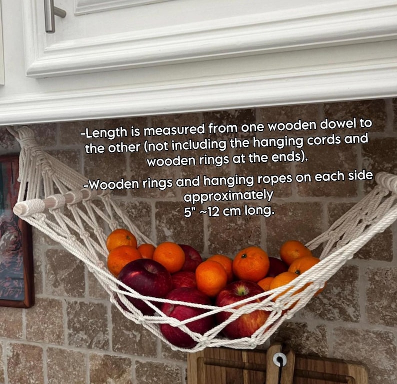May include: A white macrame fruit hammock, filled with red apples and oranges, suspended from a wooden dowel. The text on the image says the wooden rings and hanging ropes are approximately 12 cm long. The hammock is made of white rope.