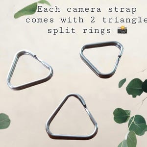 May include: Three silver-colored metal triangle split rings for camera straps. The text in the image reads: Each camera strap comes with 2 triangle split rings. These metallic components are designed for attaching straps to camera bodies.