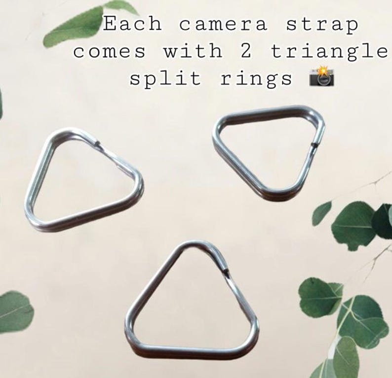 May include: Three silver-colored metal triangle split rings for camera straps. The text in the image reads: Each camera strap comes with 2 triangle split rings. These metallic components are designed for attaching straps to camera bodies.