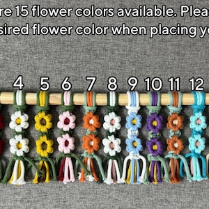 May include: Fifteen macrame flower keychains hanging from a wooden dowel, numbered 1 through 15. Each keychain features two stacked flowers with varying color combinations. Text reads: There are 15 flower colors available. Please choose your desired flower color when placing your order!
