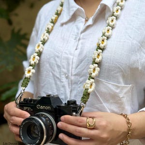 May include: A camera strap featuring a macrame design with repeating white daisy flowers with yellow centers on a dark olive green cord. The strap is attached to a black Canon FTb camera. The design consists of knotted cord forming floral shapes.