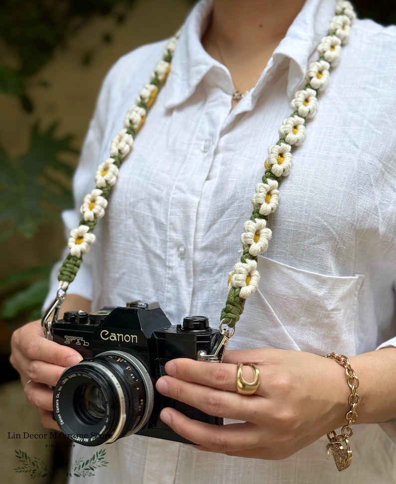 May include: A camera strap featuring a macrame design with repeating white daisy flowers with yellow centers on a dark olive green cord. The strap is attached to a black Canon FTb camera. The design consists of knotted cord forming floral shapes.
