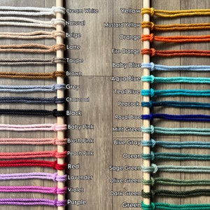 May include: A wooden dowel rod with 32 different colors of macrame cord wrapped around it. The colors are labeled with their names, including Cream White, Yellow, Natural, Mustard Yellow, Beige, Orange, Latte, Fire Orange, Taupe, Baby Blue, Brown, Aqua Blue, Grey, Teal Blue, Charcoal, Peacock, Black, Royal Blue, Baby Pink, Mint Green, Blush Pink, Blue Gray, Peach Pink, Ocean, Red, Sage Green, Lavender, Olive Green, Violet, Dark Green, Purple, and Green. The text "LINDECORMACRAMESTORE.ETSY.COM" is at the bottom of the image.