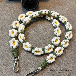 May include: A camera strap featuring a series of crocheted white daisy flowers with yellow centers, connected by a green cord. The strap includes two silver-toned metal swivel lobster clasps at the ends. Text reads Lin Decor Macrame Store.
