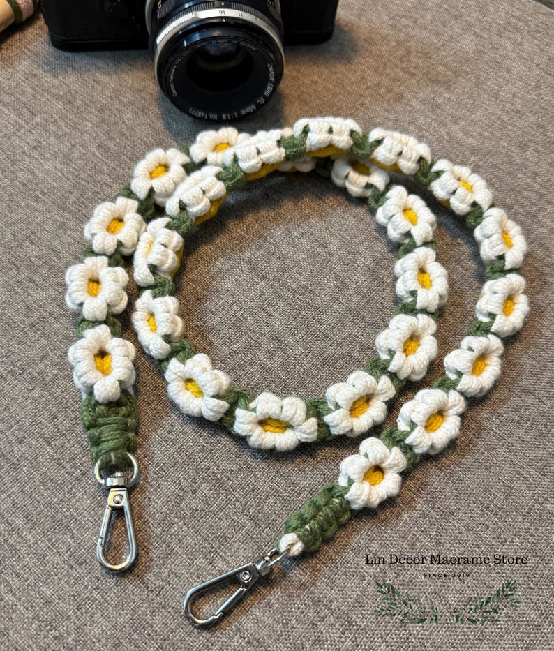 May include: A camera strap featuring a series of crocheted white daisy flowers with yellow centers, connected by a green cord. The strap includes two silver-toned metal swivel lobster clasps at the ends. Text reads Lin Decor Macrame Store.