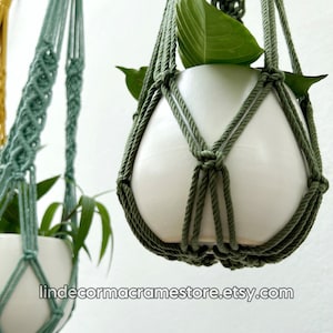May include: Close-up of two macrame plant hangers in shades of green. Each hanger cradles a white ceramic pot with green leafy plants. The macrame design is visible, with the text "lindecormacramestore.etsy.com" at the bottom.