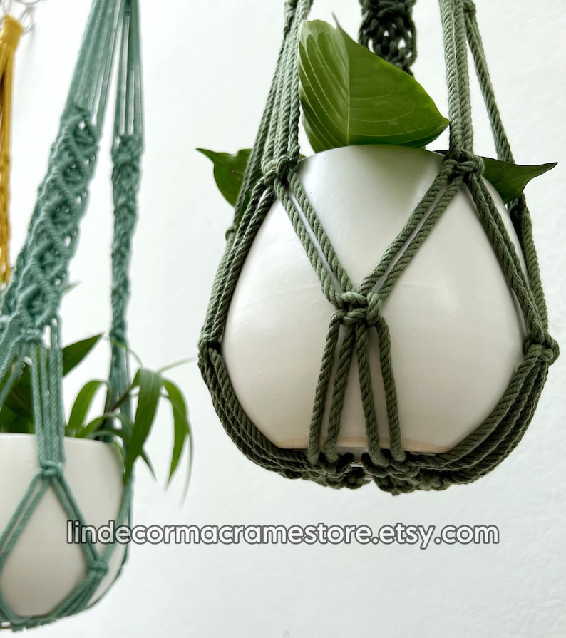 May include: Close-up of two macrame plant hangers in shades of green. Each hanger cradles a white ceramic pot with green leafy plants. The macrame design is visible, with the text "lindecormacramestore.etsy.com" at the bottom.