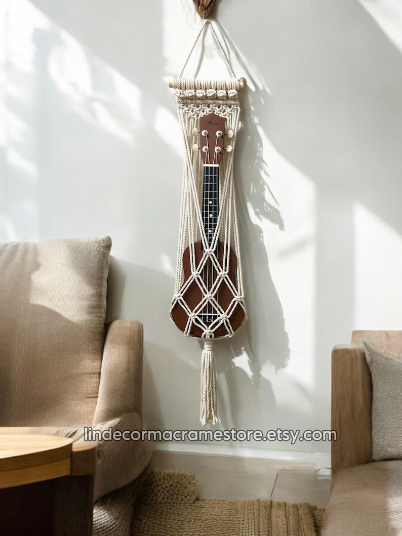 May include: A white macrame wall hanging with a wooden dowel at the top, designed to hold a ukulele. The ukulele is brown with white tuning pegs and is secured in the macrame net. The wall hanging has a long white tassel at the bottom. The wall hanging is displayed on a white wall with a brown couch and a wooden table in the foreground.