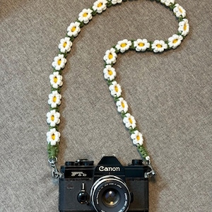 May include: A black Canon FTb camera attached to a handmade macrame camera strap featuring a repeating pattern of white and yellow daisy flowers on a green cord. The text Lin Decor Macrame Store SINCE 2019 is printed in the bottom left corner.