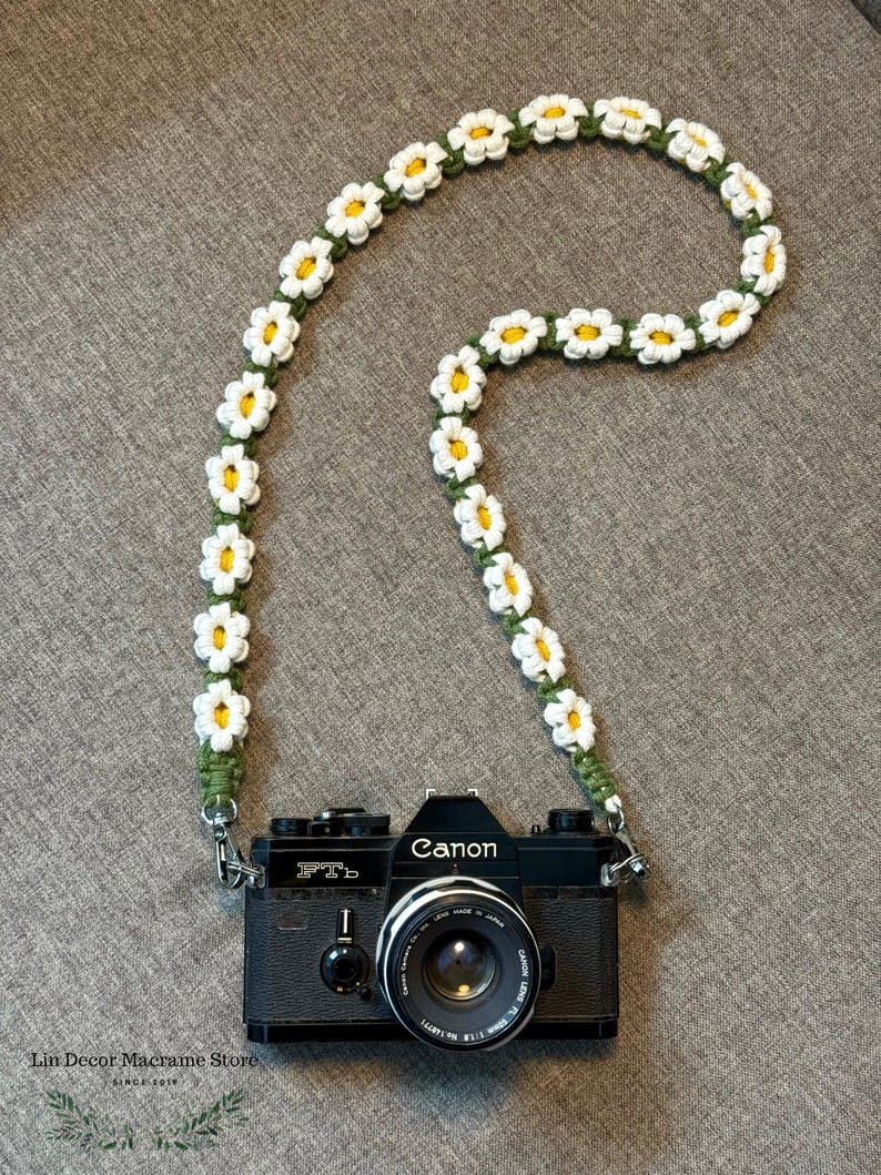 May include: A black Canon FTb camera attached to a handmade macrame camera strap featuring a repeating pattern of white and yellow daisy flowers on a green cord. The text Lin Decor Macrame Store SINCE 2019 is printed in the bottom left corner.
