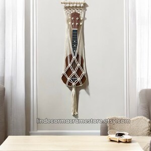 May include: A white macrame wall hanging with a wooden dowel at the top. The hanging is designed to hold a ukulele, which is visible in the center of the hanging. The hanging has a long tassel at the bottom. The hanging is displayed on a white wall with a wooden table and a chair in the background. The text "lindecormacramestore.etsy.com" is visible at the bottom of the image.