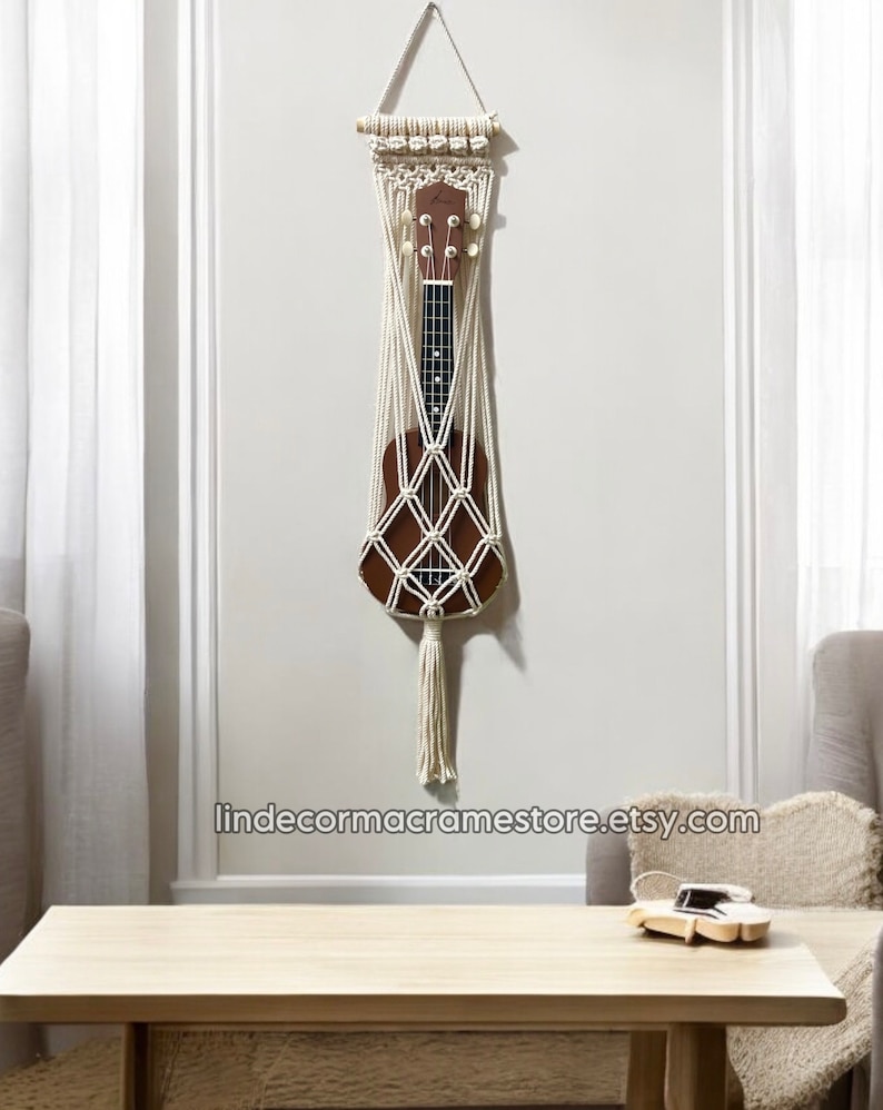 May include: A white macrame wall hanging with a wooden dowel at the top. The hanging is designed to hold a ukulele, which is visible in the center of the hanging. The hanging has a long tassel at the bottom. The hanging is displayed on a white wall with a wooden table and a chair in the background. The text "lindecormacramestore.etsy.com" is visible at the bottom of the image.