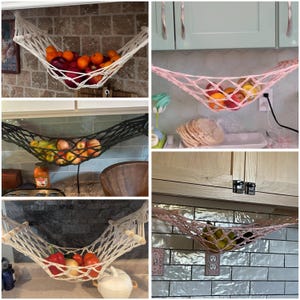 May include: Several macrame fruit hammocks in white, pink, and black, each holding fresh fruit and vegetables. The hammocks, suspended from kitchen units, offer a decorative storage solution. The produce includes oranges, apples, and tomatoes.