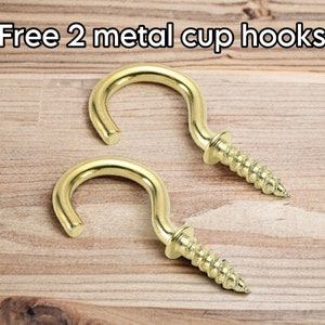 May include: Two gold-coloured metal cup hooks with screw threads, displayed on a light brown wooden surface. The text "Free 2 metal cup hooks" is at the top of the image.