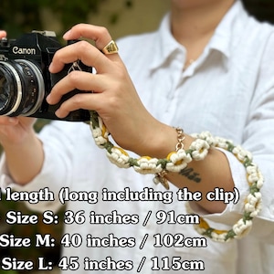 May include: A camera strap featuring a repeating pattern of crocheted white and yellow daisy flowers with green stems. The strap is attached to a black Canon camera. Text indicates total lengths: Size S 36 inches, Size M 40 inches, Size L 45 inches.