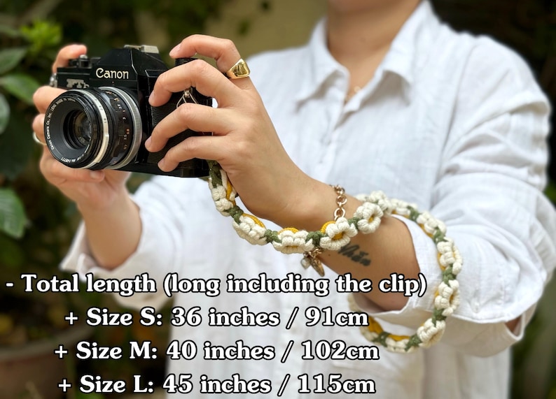 May include: A camera strap featuring a repeating pattern of crocheted white and yellow daisy flowers with green stems. The strap is attached to a black Canon camera. Text indicates total lengths: Size S 36 inches, Size M 40 inches, Size L 45 inches.
