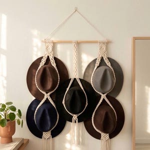 May include: A wall-mounted macrame hat organizer featuring a light-colored wooden dowel and off-white knotted cord. The organizer holds five wide-brimmed felt hats in shades of brown, black, gray, and navy, arranged in a tiered, symmetrical pattern.