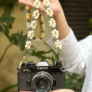 May include: A black Canon FTb film camera with a lens attached, featuring a handmade crochet camera strap. The strap is constructed from a series of white daisy-shaped flowers with yellow centers, connected by olive green macrame cord.