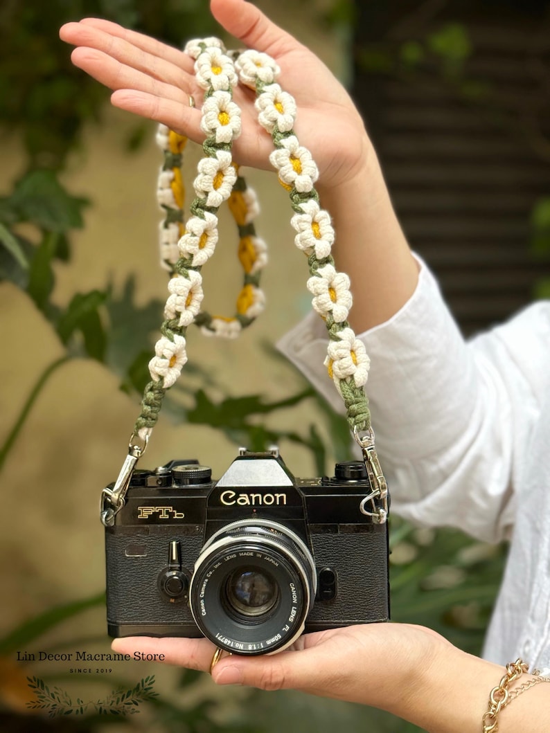 May include: A black Canon FTb film camera with a lens attached, featuring a handmade crochet camera strap. The strap is constructed from a series of white daisy-shaped flowers with yellow centers, connected by olive green macrame cord.