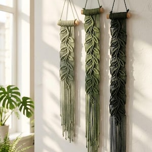 May include: Three vertical macrame wall hangings suspended from wooden dowels. Each piece features a knotted leaf pattern in varying shades of green, ranging from light sage to dark forest green, with long, loose fringe ends at the bottom.