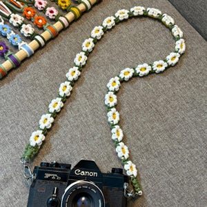 May include: A black Canon FTb camera attached to a long, handmade crochet camera strap featuring a repeating pattern of white daisy flowers with yellow centers on a green cord. The strap is connected to the camera via silver metal clips.