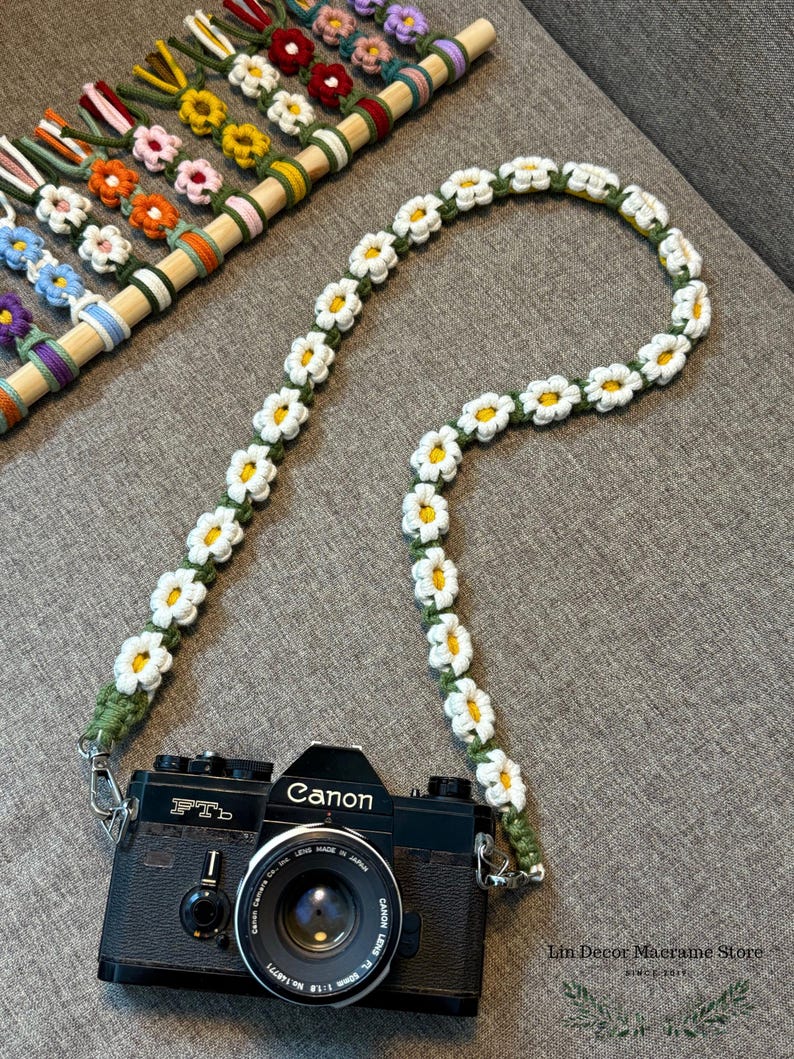 May include: A black Canon FTb camera attached to a long, handmade crochet camera strap featuring a repeating pattern of white daisy flowers with yellow centers on a green cord. The strap is connected to the camera via silver metal clips.