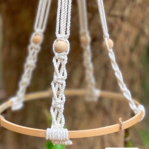 May include: A white macrame plant hanger with a wooden ring and wooden beads. The hanger is made with a thick, natural cotton rope.