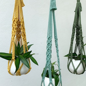 May include: Three macrame plant hangers in mustard yellow, seafoam green, and olive green, each holding a white ceramic pot with green plants. The hangers are suspended against a white wall. The Etsy shop name "lindecormacramestore.etsy.com" is visible at the bottom.